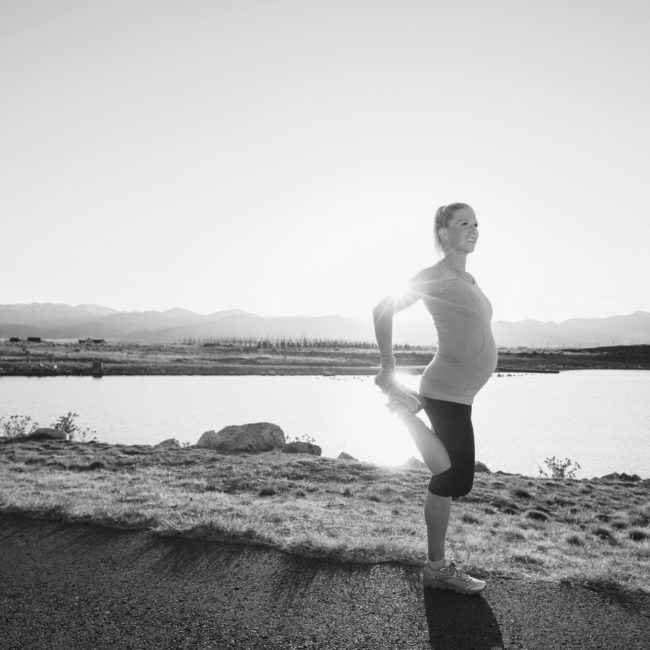 A pregnant female runner in a blue athletic shirt and running shoes is training while pregnant for her next race. She warms up by stretching with the sun flaring behind her. This woman enjoys running on trails and at sunset in the evening. Running is her favorite exercise to stay fit and happy.